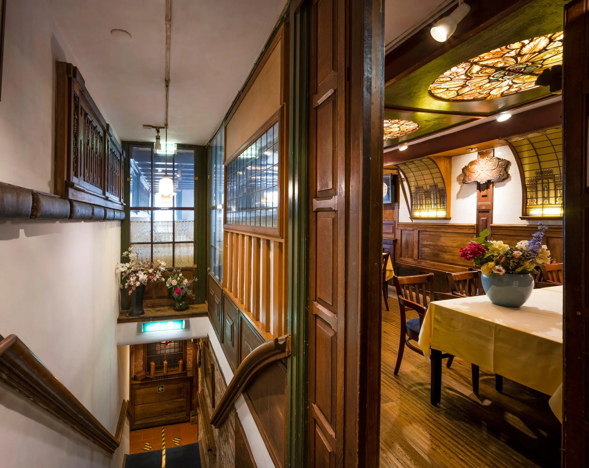 Historisch interieur met houten lambrisering, glas-in-loodramen en een trap die leidt naar een lager gelegen gang op de Nieuwezijds Voorburgwal.